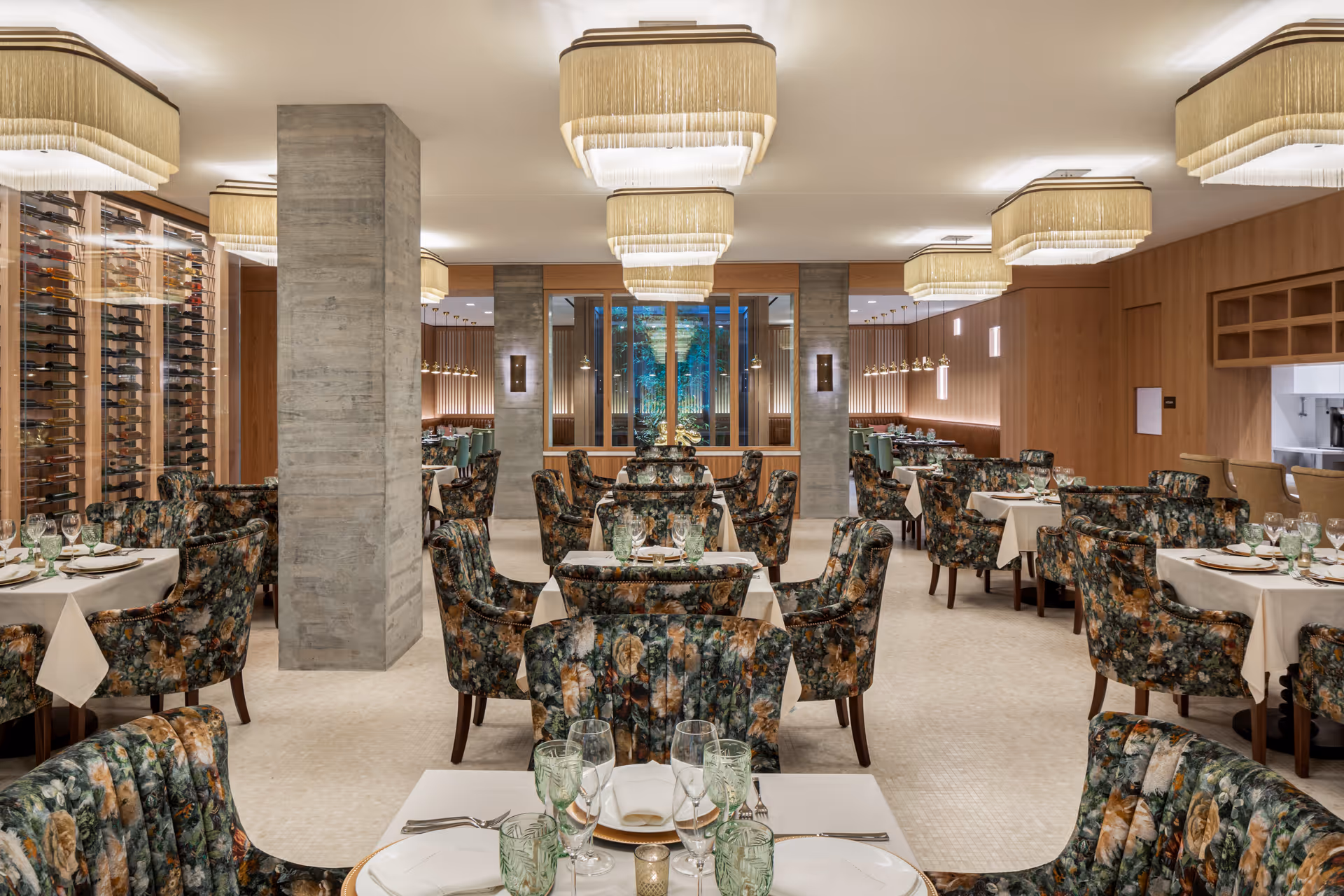 A spacious, elegant dining room with multiple tables set with white tablecloths, plates, glasses, and silverware. The chairs have a floral upholstery pattern. The room features large, rectangular chandeliers with fringe, concrete pillars, wooden paneling on the walls, and a glass-enclosed wine display on the left side.