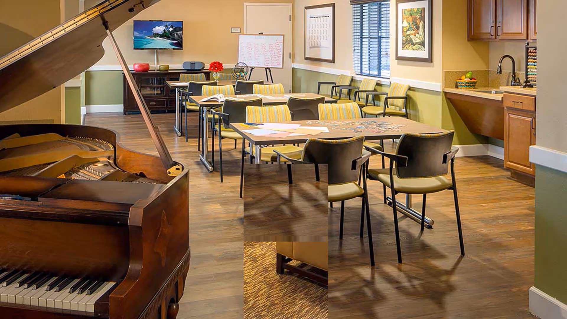 A well-lit activity room with a grand piano on the left, several tables and chairs arranged for group activities, a whiteboard with writing, a television mounted on the wall, and a kitchenette area with a sink and cabinets on the right.
