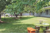 Grassy courtyard with trees, a green round outdoor table, orange benches, and a single-story building with windows.