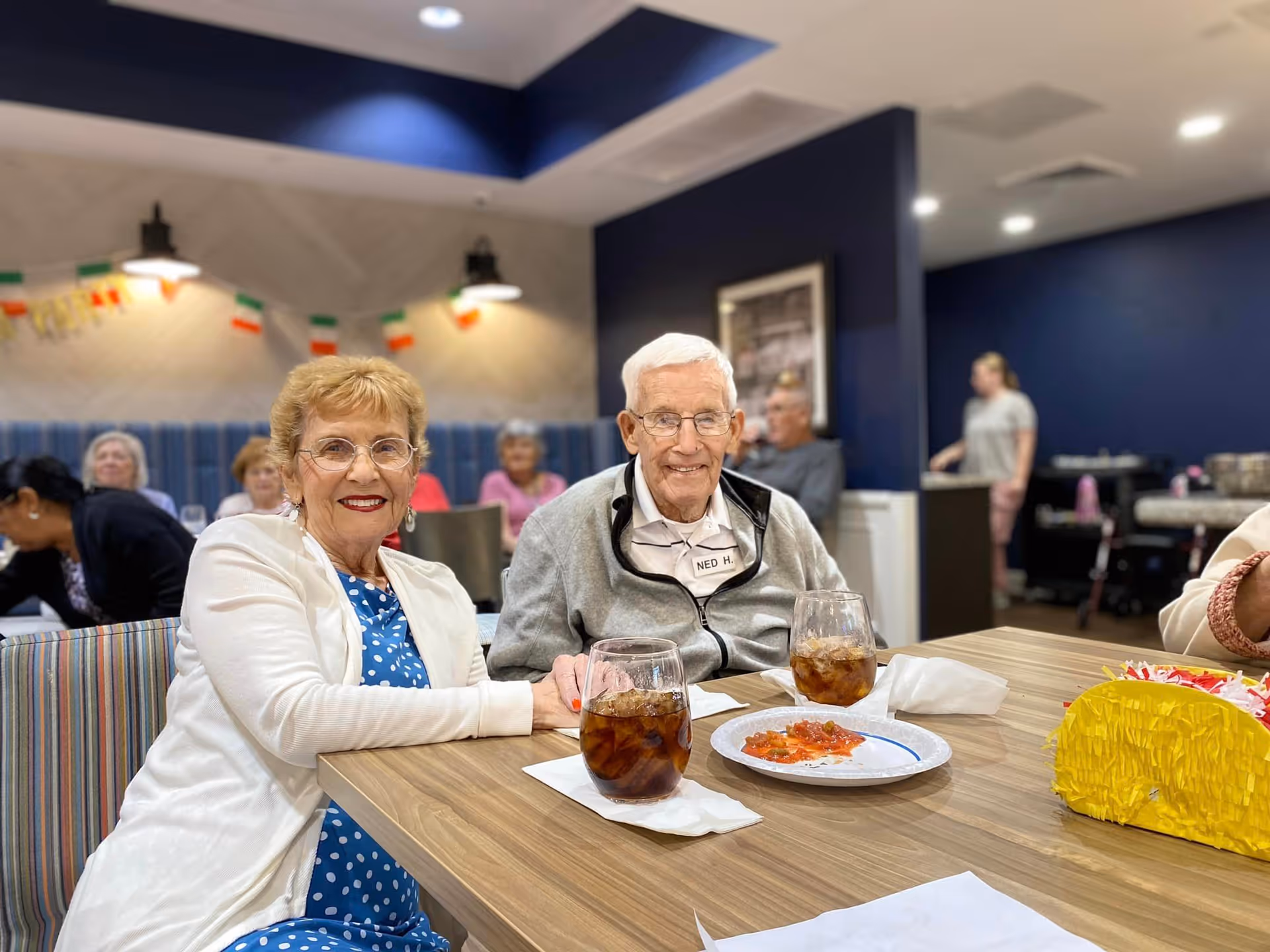 Two elderly individuals, a woman and a man, sitting at a table in a dining area. The woman is wearing glasses, a white cardigan, and a blue polka dot dress, smiling at the camera. The man, wearing glasses and a gray jacket with a name tag that reads 'Ned H.', is also smiling. On the table are two glasses of iced tea, a plate with some food, and a yellow decorative item. Other people are visible in the background, seated and standing in a well-lit room with blue walls and festive decorations.