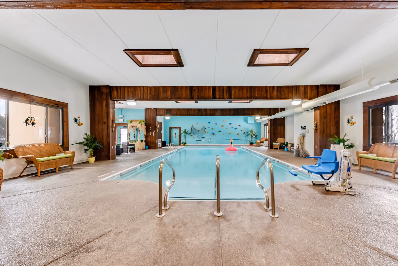 Indoor swimming pool area with a light blue pool, metal handrails, and a blue pool lift chair on the right side. The room features wooden accents, large windows, wicker seating with green cushions, tropical-themed wall decorations, and a pink flamingo float in the pool.