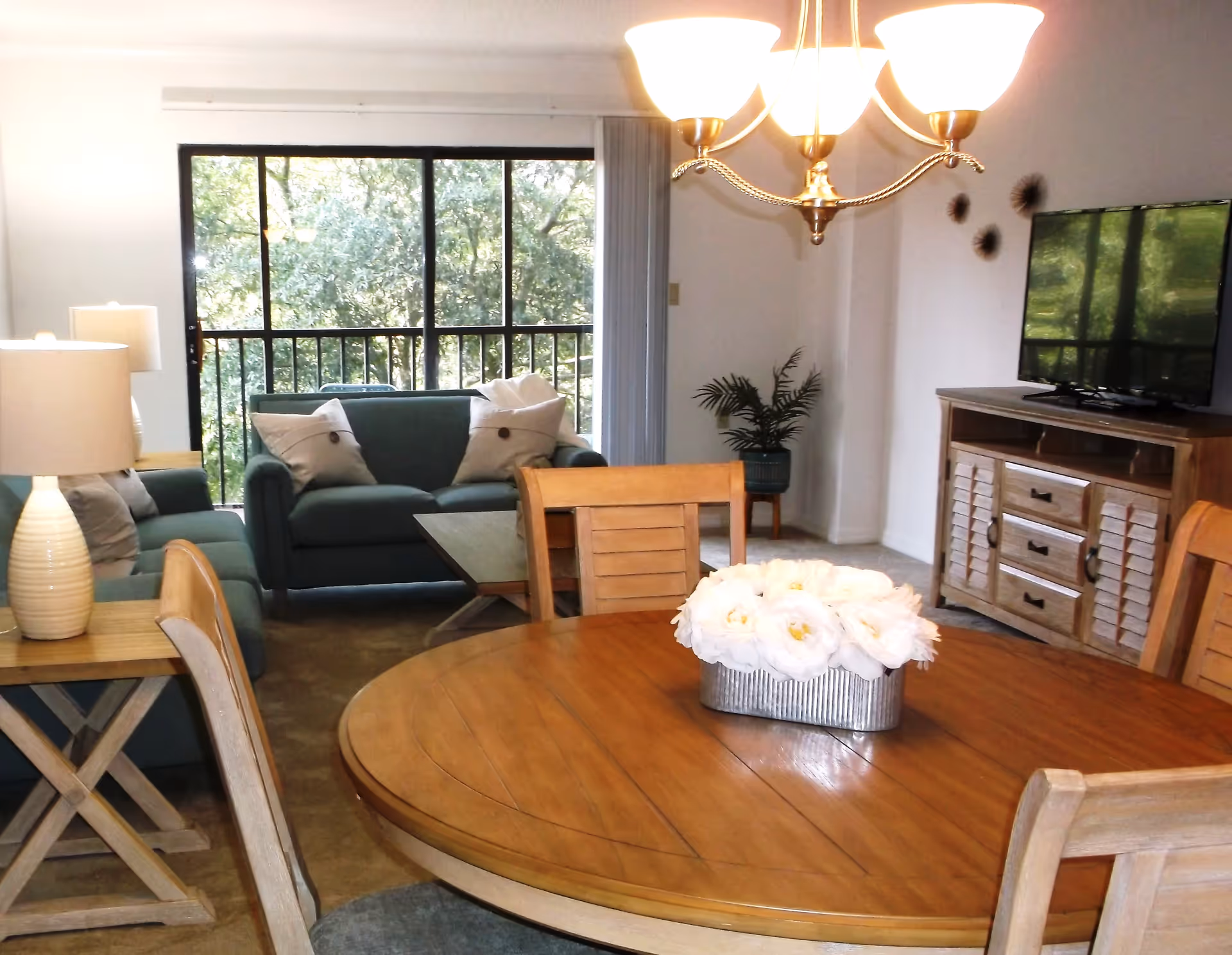 A cozy living and dining area in Hampton Apartments featuring a round wooden dining table with a floral centerpiece, wooden chairs, two teal sofas with white pillows, a wooden coffee table, a TV on a wooden stand, a potted plant in the corner, and large sliding glass doors opening to a balcony with a view of green trees.