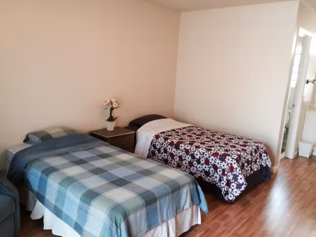 A simple bedroom with two single beds, one with a blue and green plaid bedspread and the other with a floral bedspread. There is a small wooden nightstand between the beds with a potted plant on top. The room has light-colored walls and wood flooring. A bathroom is visible through an open door on the right side of the image.