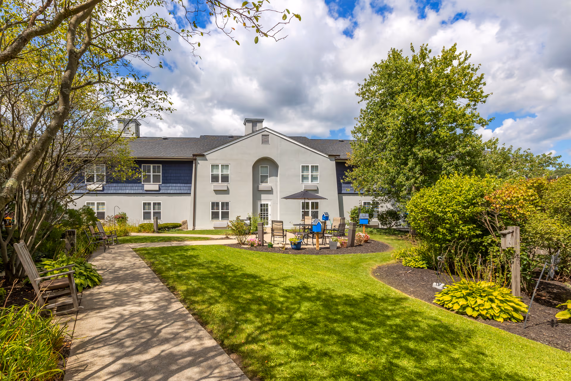 Outdoor garden area at Brookdale Manlius featuring a paved walkway with wooden benches on the left, green grass, various trees and shrubs, and a patio area with chairs and a table under an umbrella in front of a two-story building under a partly cloudy sky.