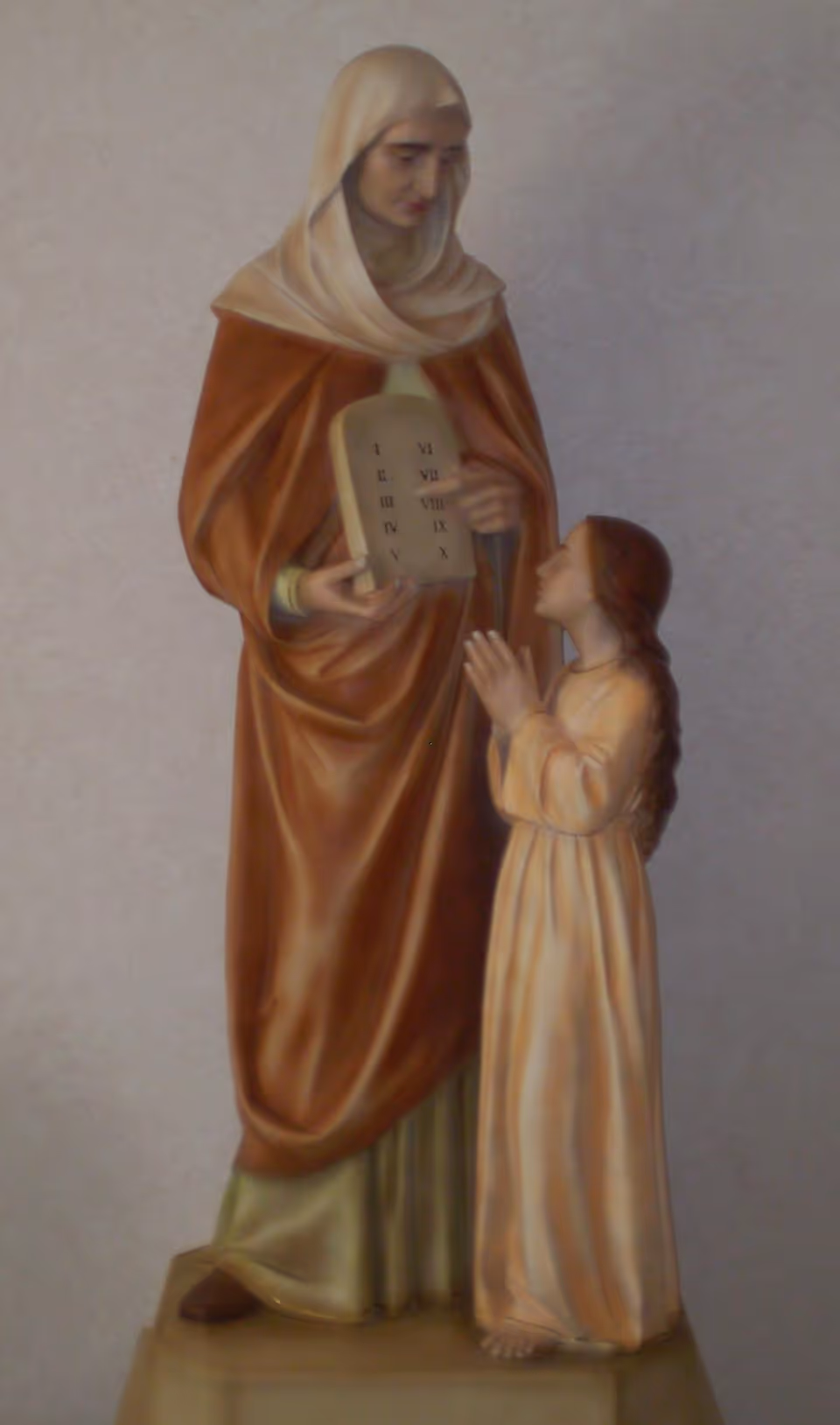 A painted statue of a robed religious figure holding tablets while a young girl stands praying beside him.