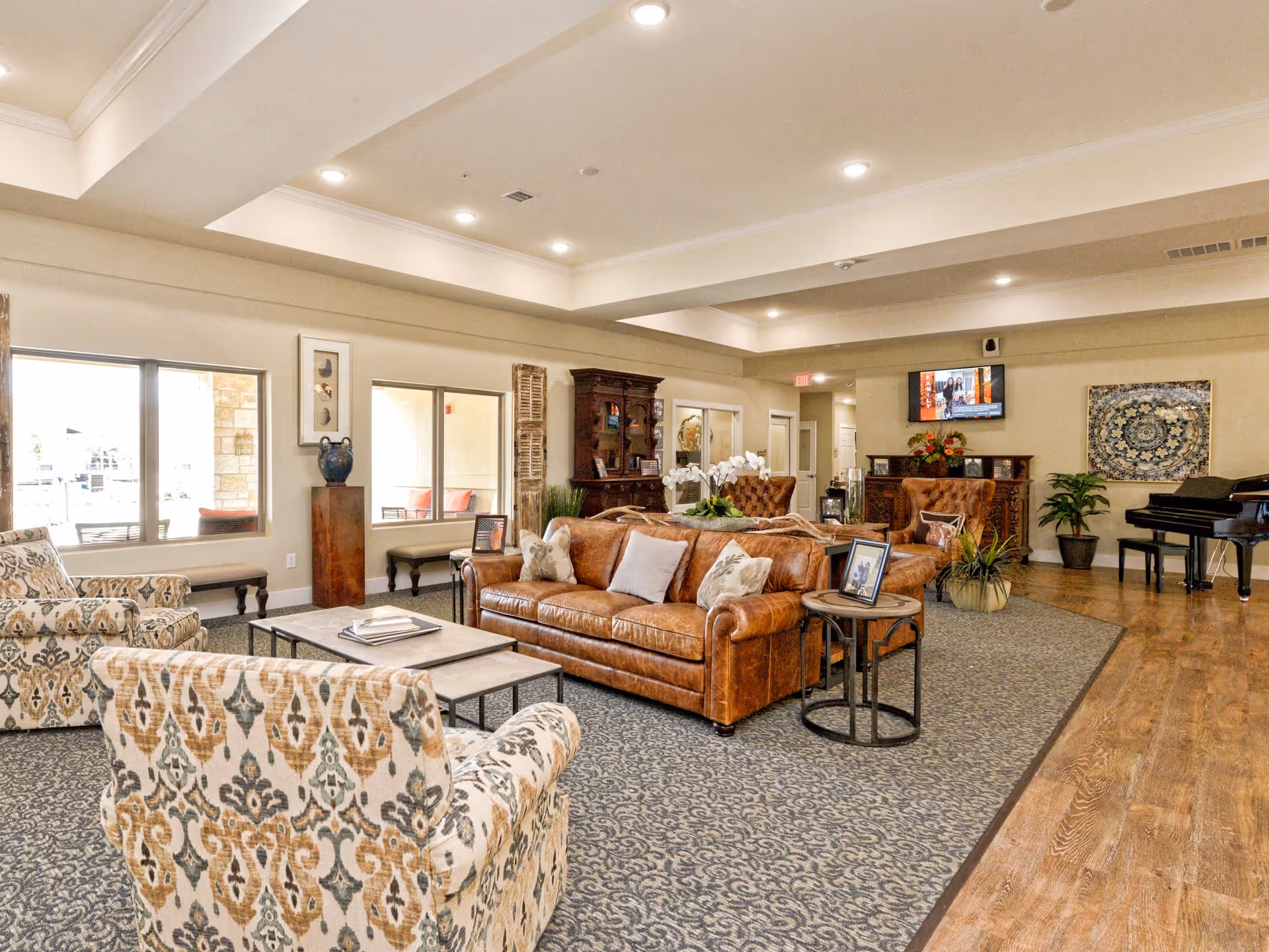 A spacious senior living facility common area with a brown leather sofa, patterned armchairs, a coffee table, and side tables. The room features a patterned carpet, wooden flooring, a grand piano, a wall-mounted TV, decorative plants, and artwork on the walls. Large windows allow natural light to enter the room.