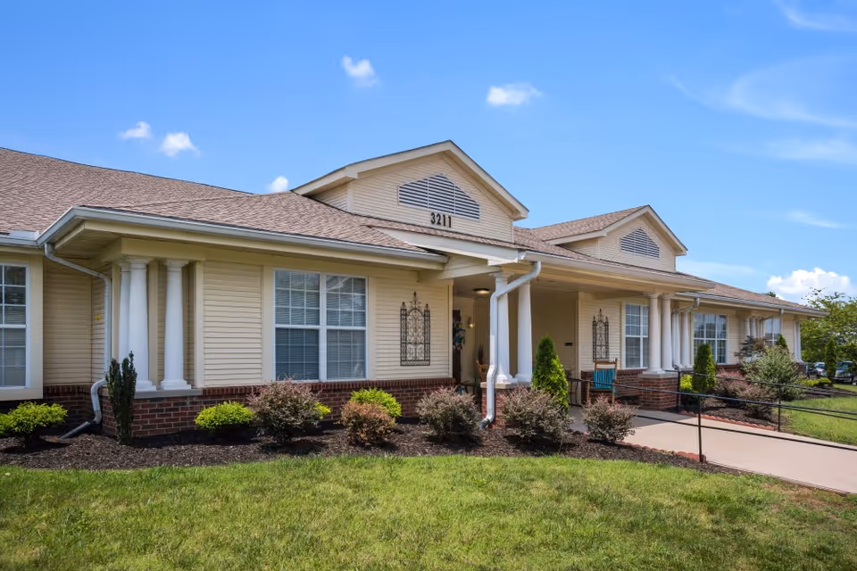 Exterior view of a single-story senior living facility building with beige siding, white columns, and a brown shingled roof under a blue sky with a few clouds. The building has large windows and a paved walkway with a handrail leading to the entrance. There are shrubs and a well-maintained lawn in front of the building.
