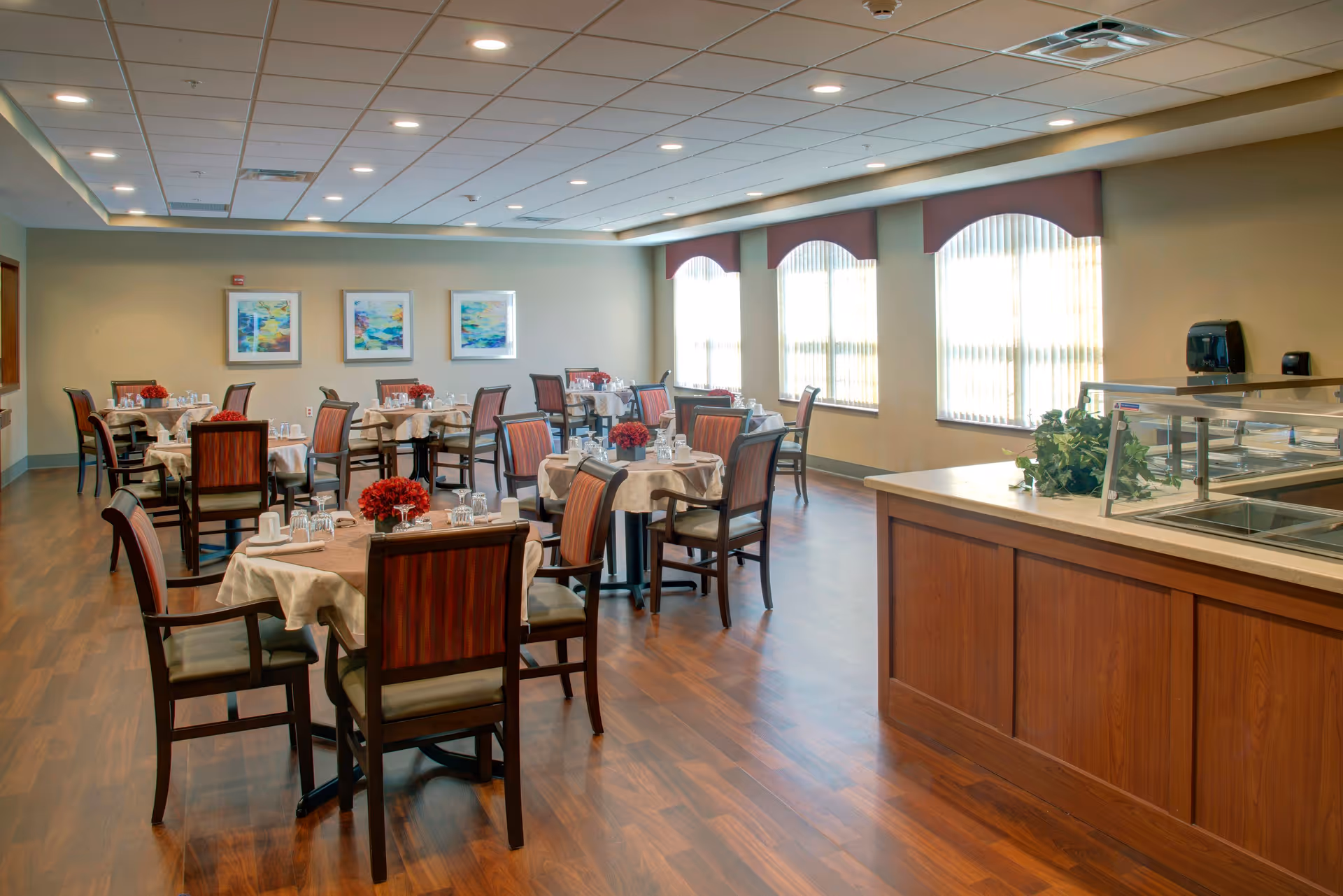 A bright dining room in a senior living facility with multiple tables covered in beige tablecloths, each set with glasses, cups, and red floral centerpieces. The room has large windows with vertical blinds, wooden flooring, and a serving counter with a glass sneeze guard and a small plant. Three colorful abstract paintings hang on the far wall.