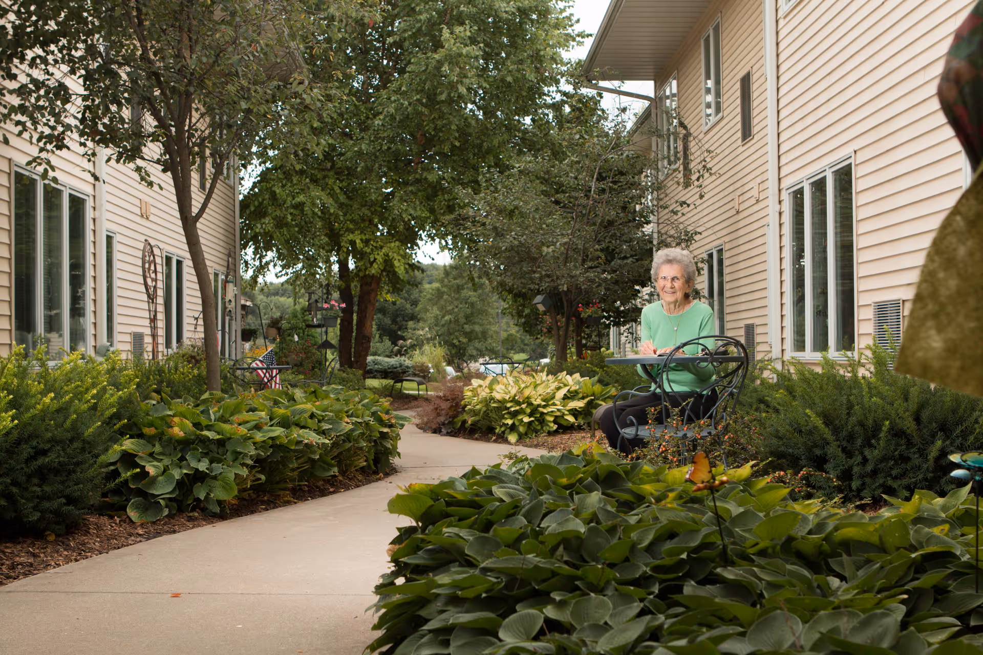 An elderly woman wearing a green sweater sits at a small outdoor table with a chair in a garden area between two beige buildings. The garden is lush with green plants, bushes, and trees lining a concrete pathway.