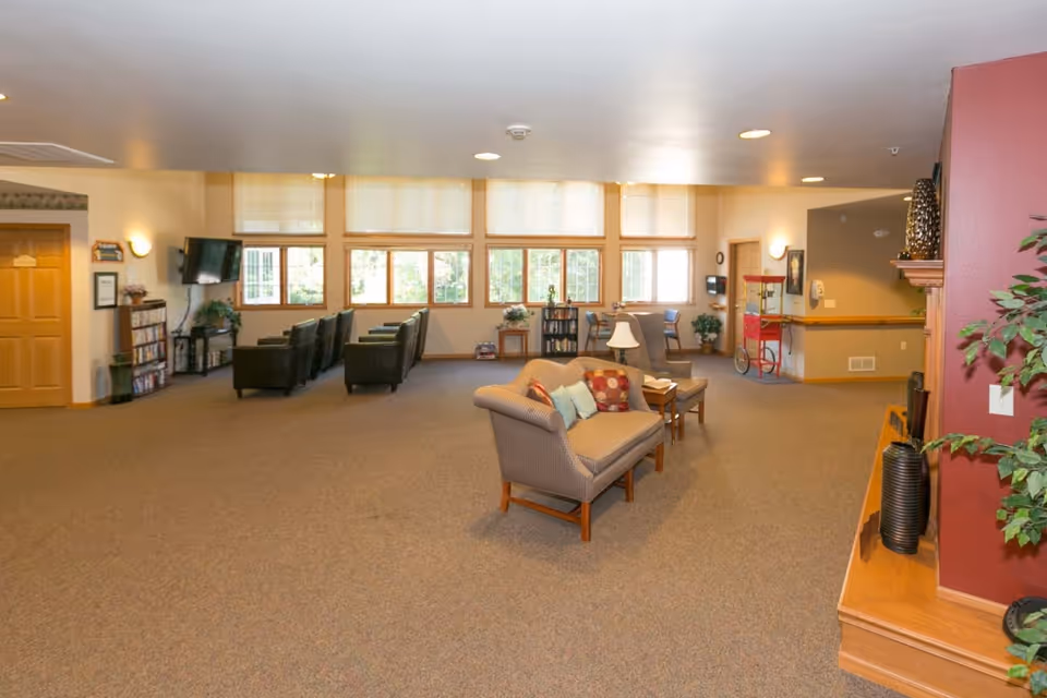 A spacious senior living common area with carpeted floor, multiple seating arrangements including armchairs and a loveseat with colorful pillows, large windows letting in natural light, a wall-mounted TV, bookshelves, a popcorn machine, and decorative plants.