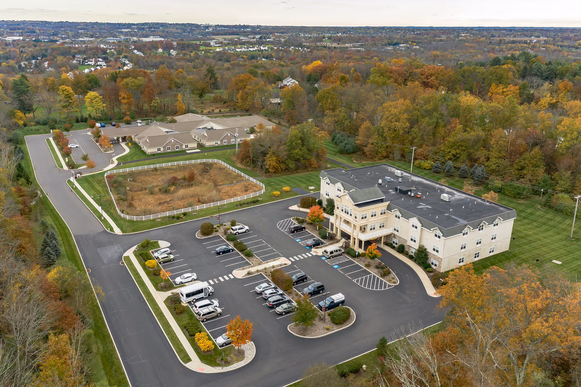 Aerial view of Hidden Meadows On the Ridge senior living facility showing a large three-story building with a parking lot in front, surrounded by trees with autumn foliage and additional smaller buildings in the background.