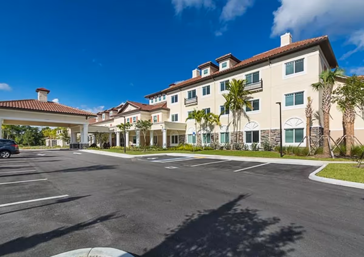 Exterior view of a multi-story senior living facility building with a covered entrance, palm trees, and a parking lot under a clear blue sky.