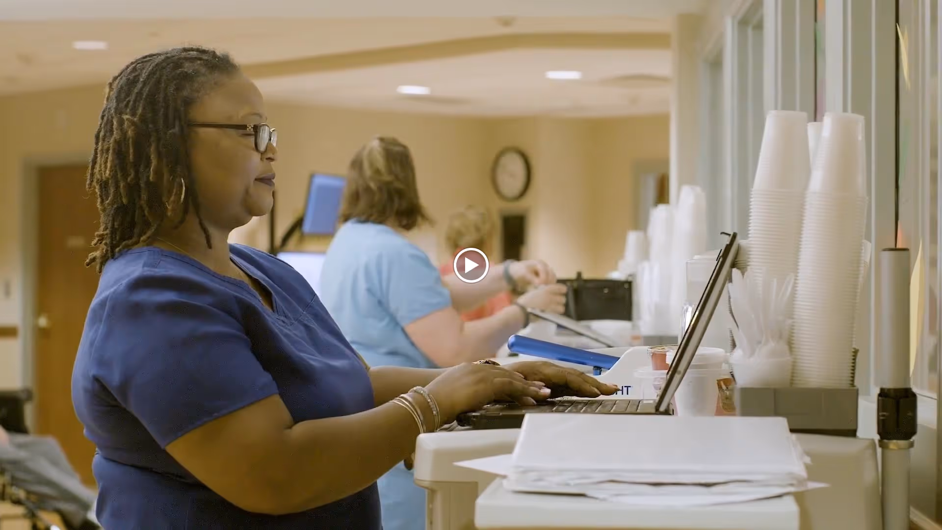 A woman in blue scrubs working on a laptop at a counter in a healthcare facility, with two other staff members in the background also engaged in tasks.