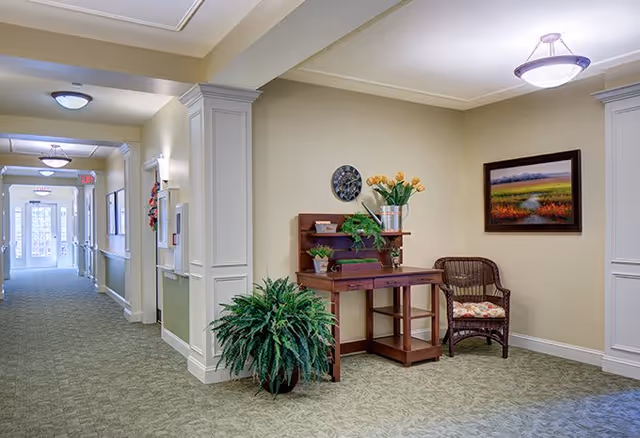 A well-lit hallway in a senior living facility with beige walls and green patterned carpet. On the right side, there is a wooden desk with plants and flowers on it, a wicker chair with a floral cushion, and a framed landscape painting on the wall. The hallway extends into the distance with multiple ceiling lights and doors along the left side.