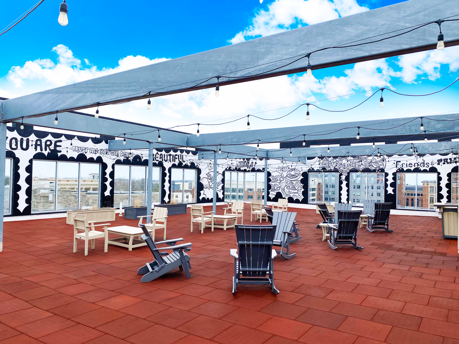 Rooftop patio with red tile flooring, scattered Adirondack chairs and tables, overhead string lights and painted mural walls overlooking city buildings.