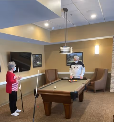Two elderly individuals wearing face masks playing pool in a well-lit room with beige walls. One person is standing near the pool table holding a cue stick, while the other is preparing to take a shot. The room has a mounted flat-screen TV, framed pictures on the wall, two armchairs, and a modern hanging light fixture above the pool table.
