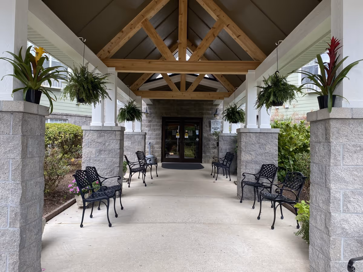 Covered entrance walkway to a building with wooden beams overhead, stone pillars on each side, black metal benches arranged along the walkway, and hanging green plants. The entrance door is glass with a sign next to it.