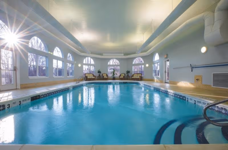 Indoor swimming pool with clear blue water surrounded by light-colored walls and large arched windows letting in natural light. There are several chairs and plants arranged along the far wall, and the pool has depth markings along the edge.