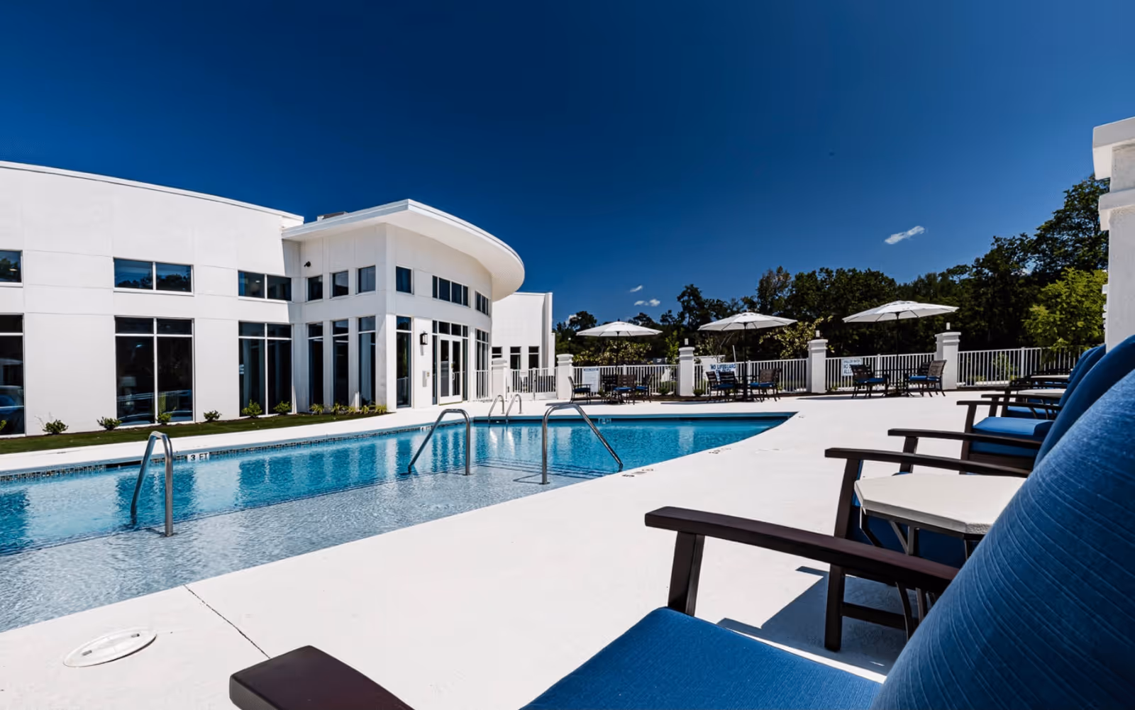 Outdoor swimming pool area at Vitality Living Columbia with blue lounge chairs in the foreground, white umbrellas, and a modern white building in the background under a clear blue sky.