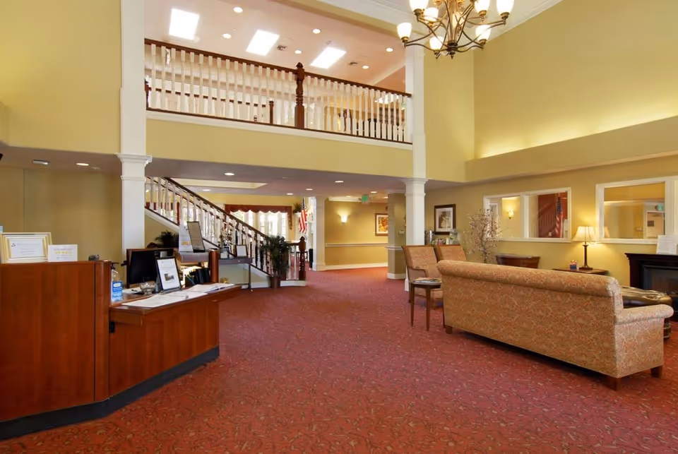 Interior view of a senior living facility lobby with a reception desk on the left, a staircase with wooden railings in the background, and a seating area with patterned sofas and chairs on the right. The space has high ceilings, yellow walls, red carpet, and warm lighting.