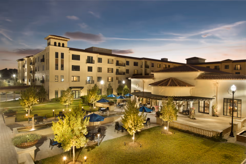 Illuminated multi-story senior living building with a landscaped courtyard, patio seating, and umbrellas at dusk.