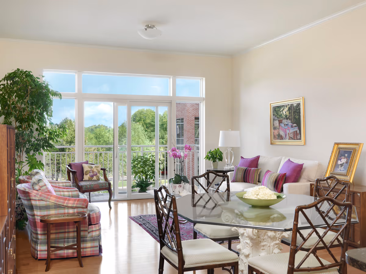 Bright and airy living room with large glass sliding doors opening to a balcony with greenery outside. The room features a glass dining table with six chairs, a white sofa with purple and striped cushions, two plaid armchairs, a wooden side table, a floor lamp, potted plants, and framed artwork on the walls.