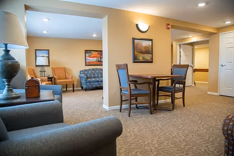 A cozy senior living common area with beige walls and carpeted floor. The room features a small round wooden table with four chairs, two armchairs in orange and blue patterns, a blue loveseat, a side table with a lamp, and framed landscape paintings on the walls. The space is well-lit with ceiling lights and a wall sconce.