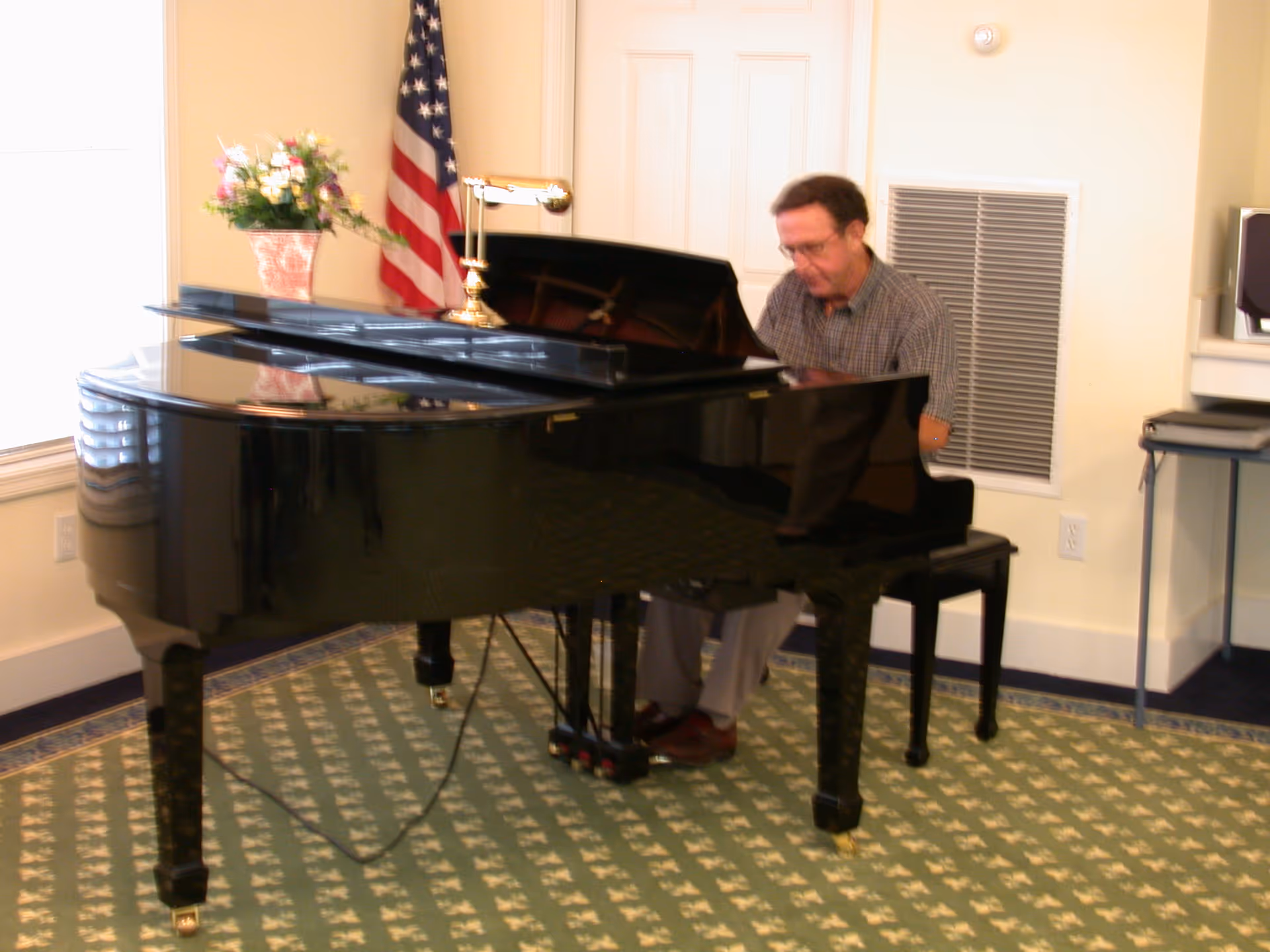 A man plays a black grand piano in a carpeted room with an American flag and a vase of flowers nearby.