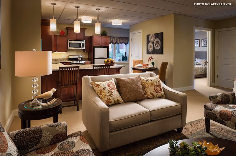 A cozy living room area with a beige loveseat adorned with patterned cushions, two patterned armchairs, a round side table with a lamp and decorative bird, and a coffee table with a plant and decorative items. In the background, there is a kitchen with dark wood cabinets, a refrigerator, microwave, stove, and a breakfast bar with three chairs. A small dining table with two chairs is visible near a window, and a bedroom can be seen through an open doorway.