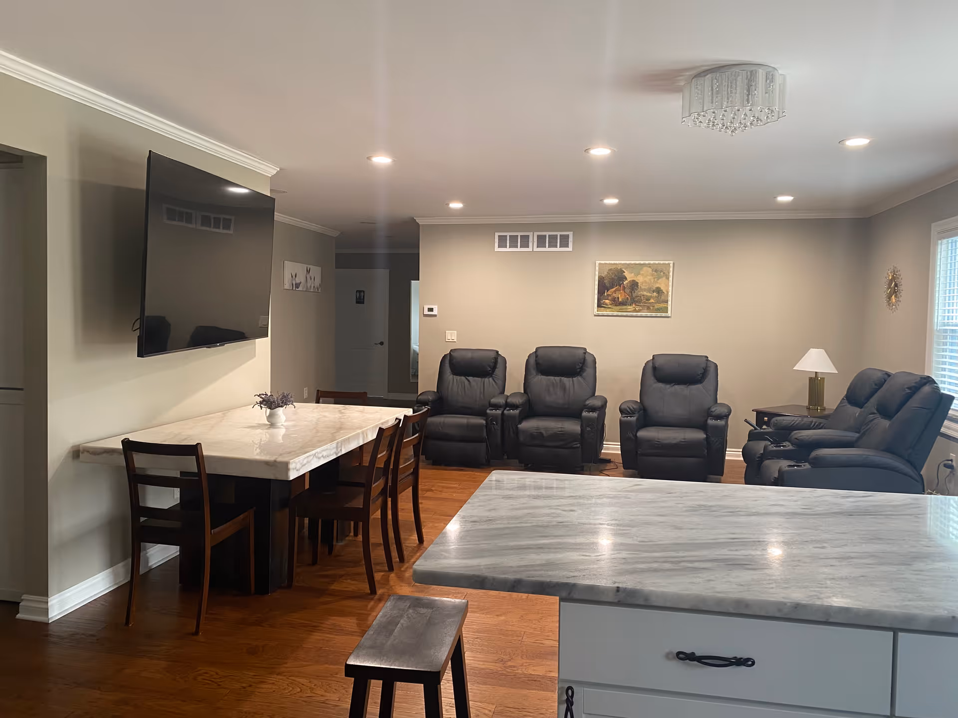 A cozy living room area with four black recliner chairs arranged against a beige wall. A framed painting hangs on the wall above the chairs. To the left, there is a dining table with four wooden chairs and a small vase with flowers. A large flat-screen TV is mounted on the wall above the dining table. The room has wooden flooring, recessed ceiling lights, a window with blinds on the right, and a marble countertop in the foreground.