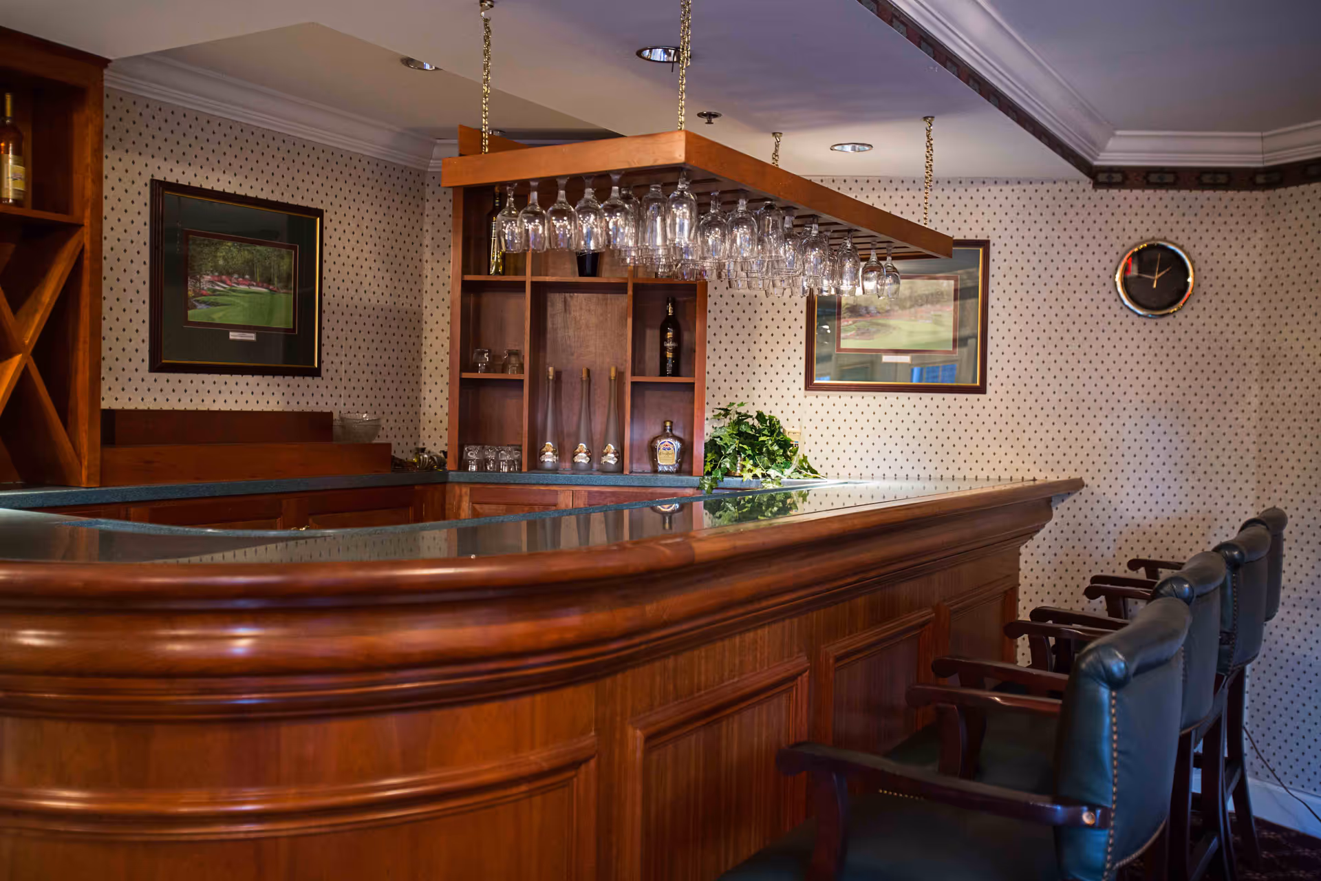Wood-paneled bar with hanging wine glasses, shelving, framed artwork and green leather bar stools in a patterned-wall interior.