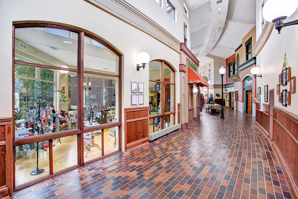 Interior hallway of a senior living facility with large windows on the left showing a gift shop area with various items including teddy bears and decorations. The hallway has a tiled floor, wood paneling on the walls, and decorative streetlamp-style lights. There is a red awning over one of the doorways and signs indicating a café further down the hall.
