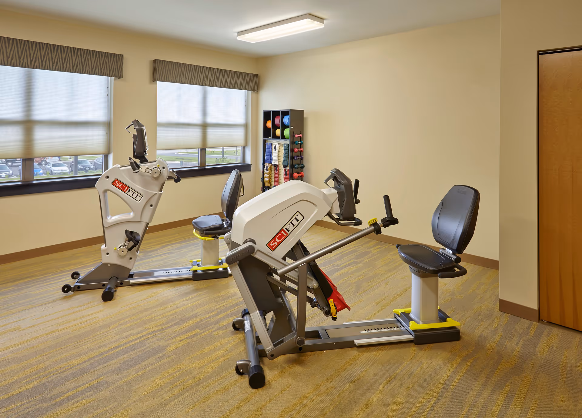 Small exercise room with two recumbent exercise machines, a rack of colorful weights and medicine balls, and windows with shades.
