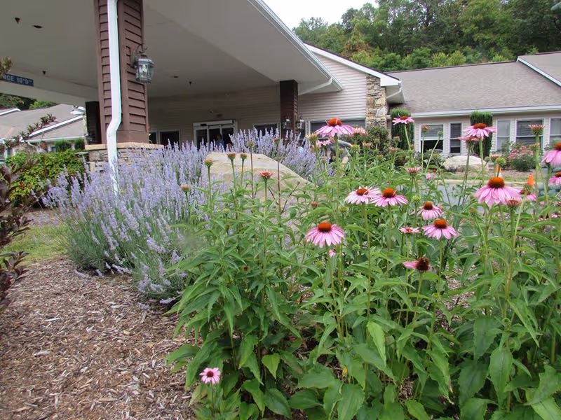 Entrance of Heather Glen At Ardenwoods facility with a covered drop-off area, surrounded by landscaped garden beds featuring purple and pink flowers, green shrubs, and a stone wall section on the building exterior.