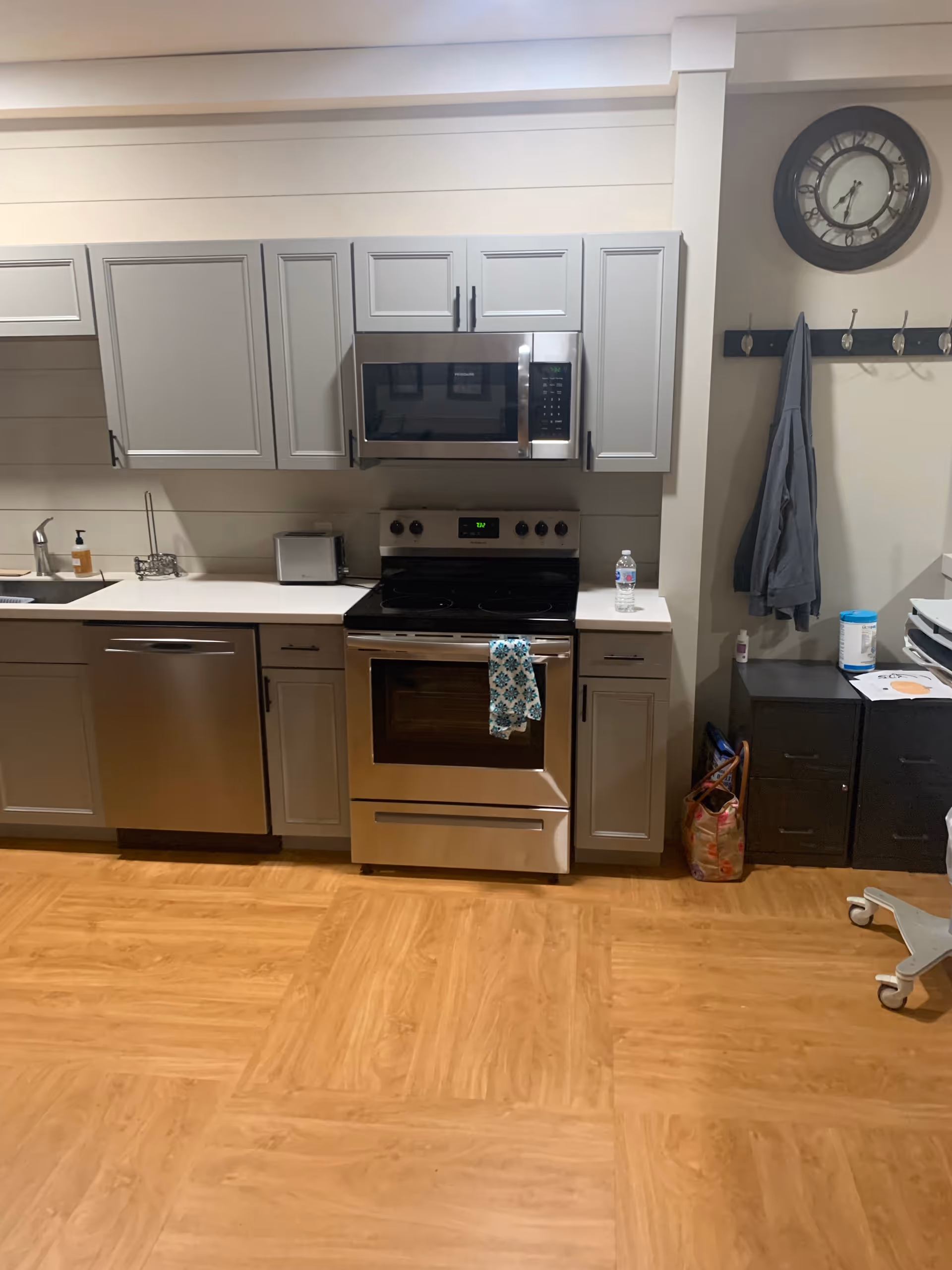 Modern kitchen area with light gray cabinets, stainless steel appliances including a microwave, stove, and dishwasher. A patterned towel hangs on the oven handle. There is a water bottle on the counter, a clock on the wall, and a coat hanging on hooks next to a small black filing cabinet and a bag on the floor. The floor is wood-patterned.