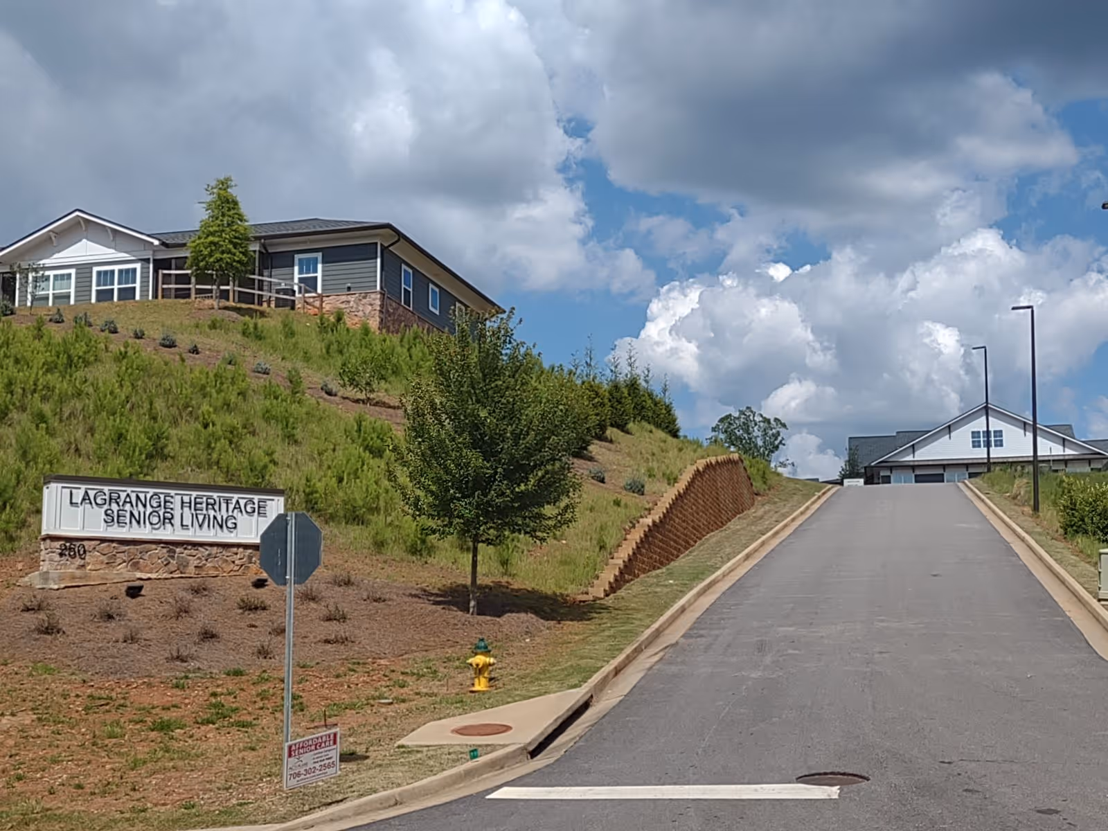 Exterior view of LaGrange Heritage Senior Living facility showing a paved driveway leading uphill towards buildings with a partly cloudy sky. There is a sign on the left side of the driveway that reads 'LAGRANGE HERITAGE SENIOR LIVING' with some landscaping and a small tree nearby.