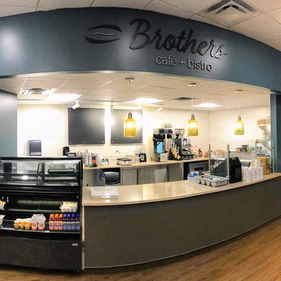 Interior view of Brothers café + bistro with a curved counter, display case with beverages and desserts, coffee machines, and hanging pendant lights.