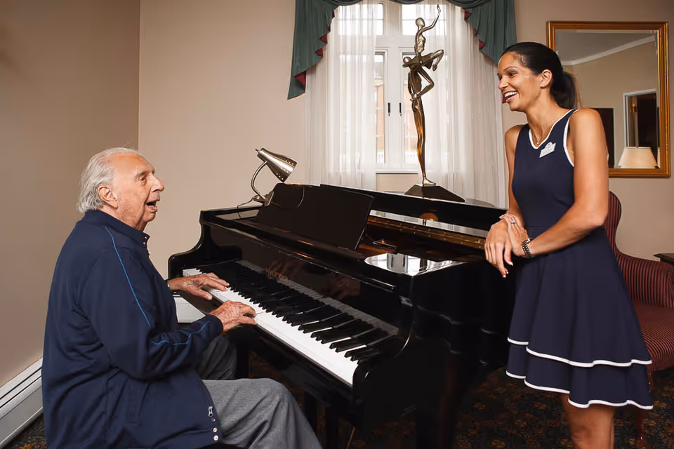 An elderly man playing a black grand piano while a woman in a navy blue dress with white trim stands nearby smiling and listening. The room has a window with sheer white curtains and green valances, a bronze sculpture on the piano, and a framed mirror on the wall.
