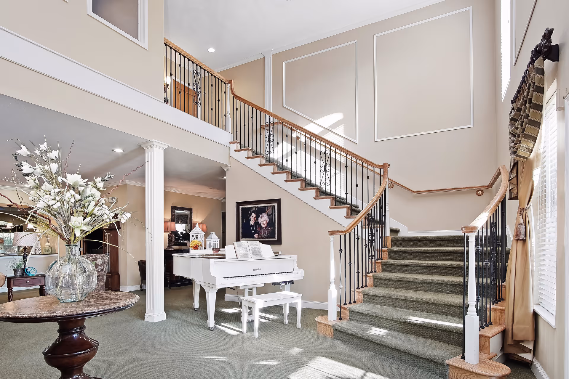 A spacious and elegant interior area featuring a carpeted staircase with wooden handrails and black iron balusters. A white grand piano with a matching bench is positioned near the staircase, with sheet music on the piano. A round wooden table with a large glass vase holding white flowers is in the foreground. The walls are painted beige with decorative molding, and large windows with curtains allow natural light to fill the space.