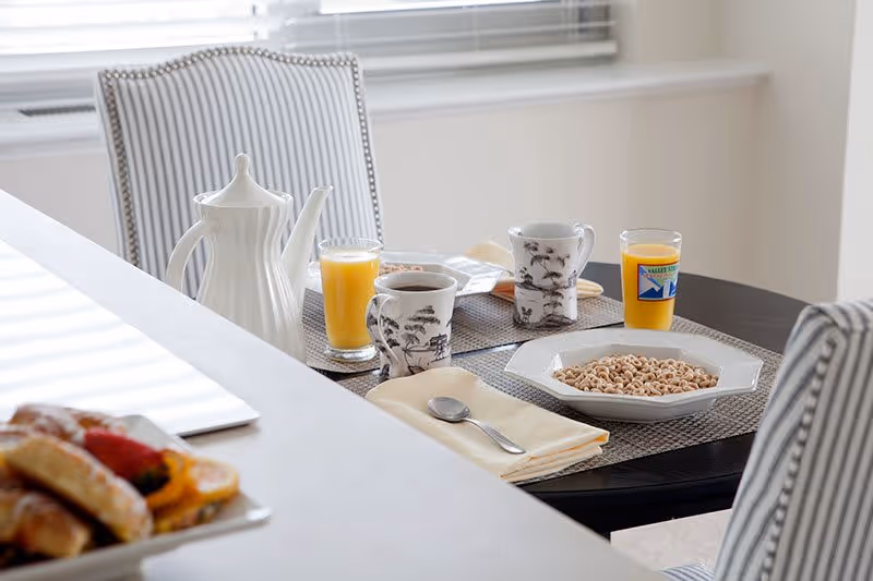 A dining table set for breakfast with two bowls of cereal, two glasses of orange juice, two mugs with a black and white tree design, a white teapot, and a plate of pastries in the foreground. The table has placemats and napkins, and there are striped upholstered chairs around it. A window with blinds is in the background.