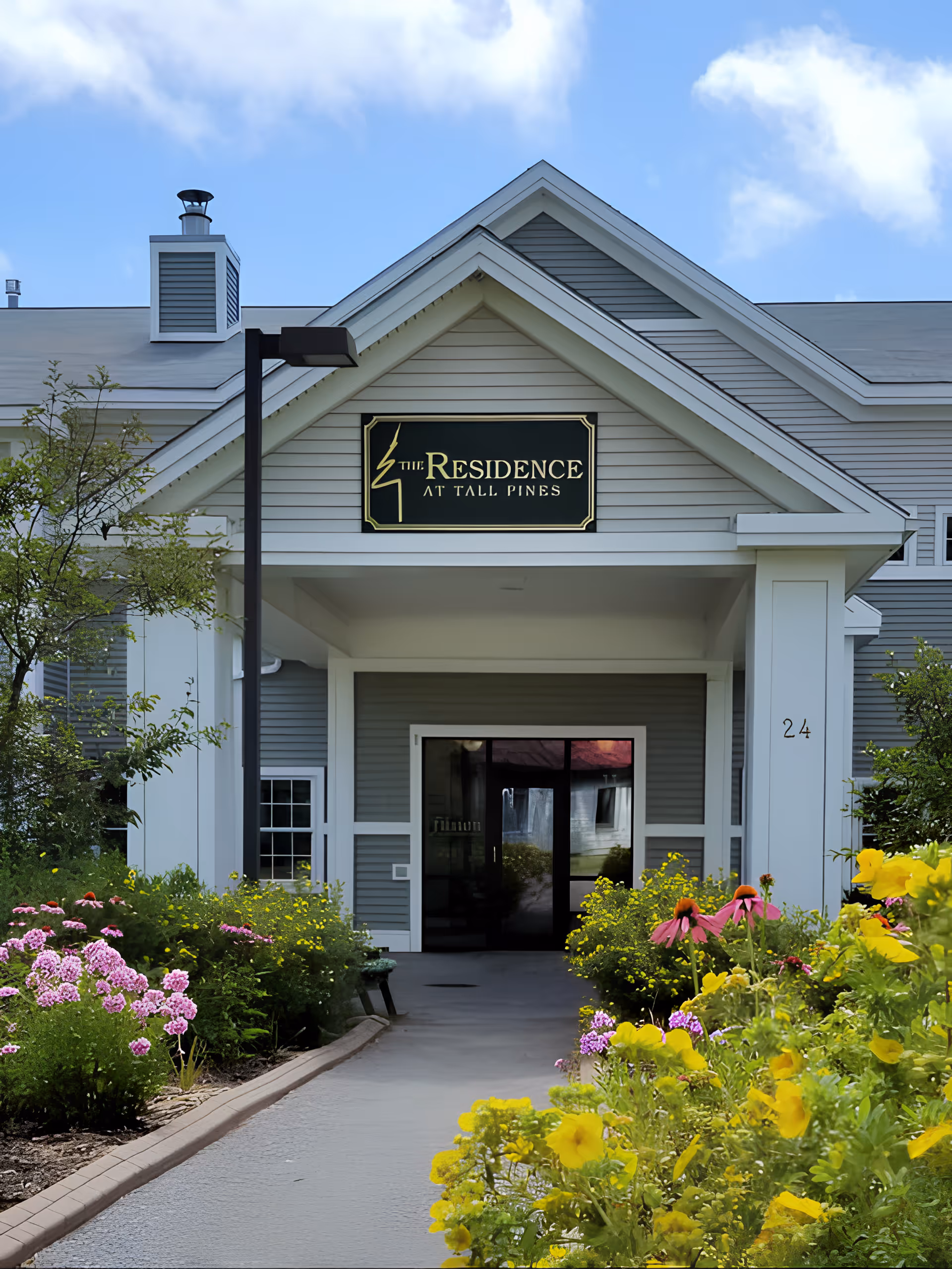 Entrance to The Residence at Tall Pines building with a covered walkway, surrounded by colorful flowers and greenery under a partly cloudy sky.