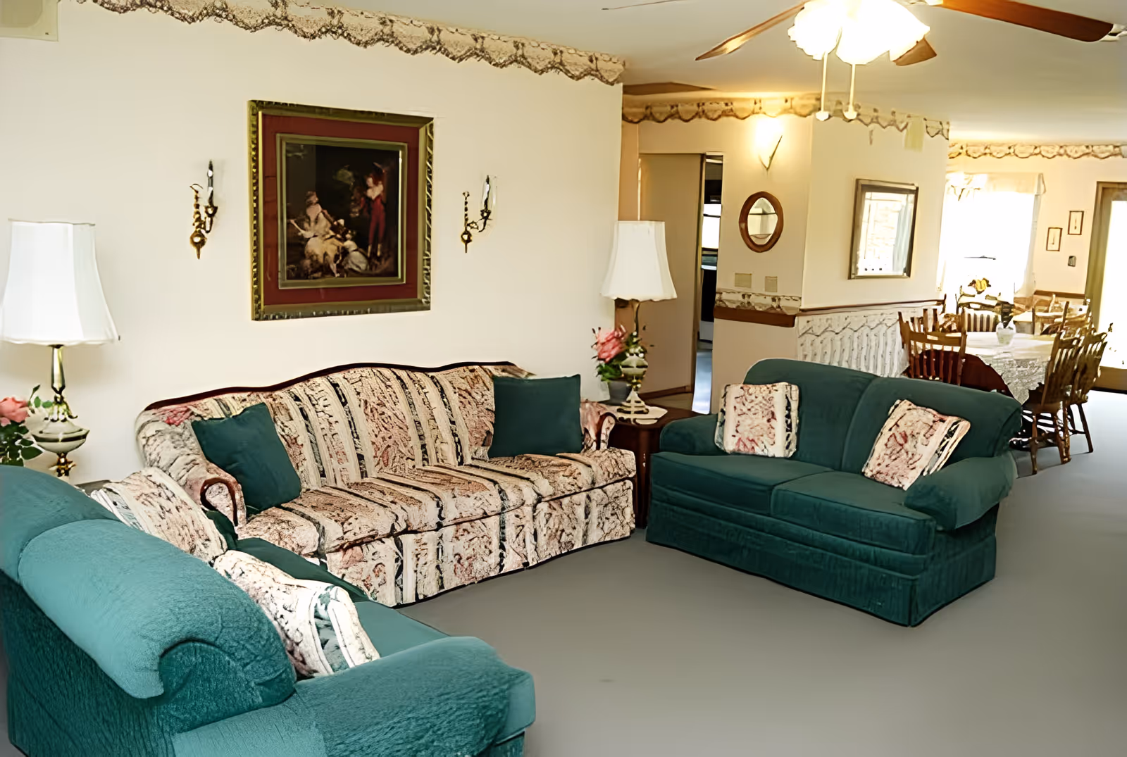 A cozy living room with two green upholstered sofas and one patterned sofa arranged around a small wooden side table with a lamp and flower vase. The walls are decorated with a framed painting and two wall sconces. In the background, a dining area with a wooden dining table and chairs is visible, illuminated by natural light from a window.