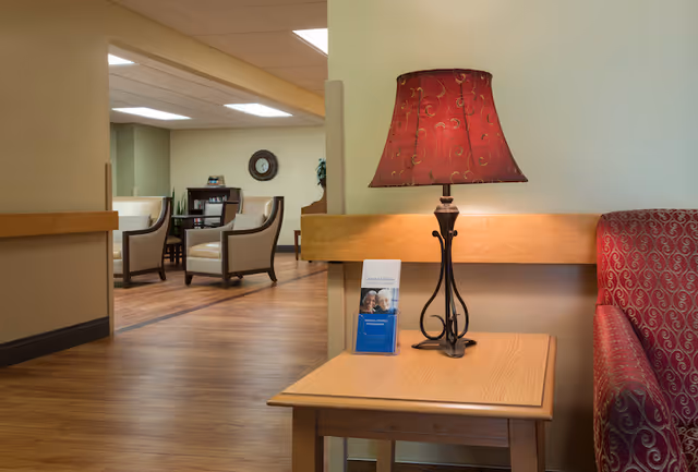 A cozy interior common area in a senior living facility with a wooden side table holding a decorative lamp with a red lampshade and a brochure. In the background, there are cushioned armchairs, a small table, a clock on the wall, and a bookshelf, all set on a wooden floor.