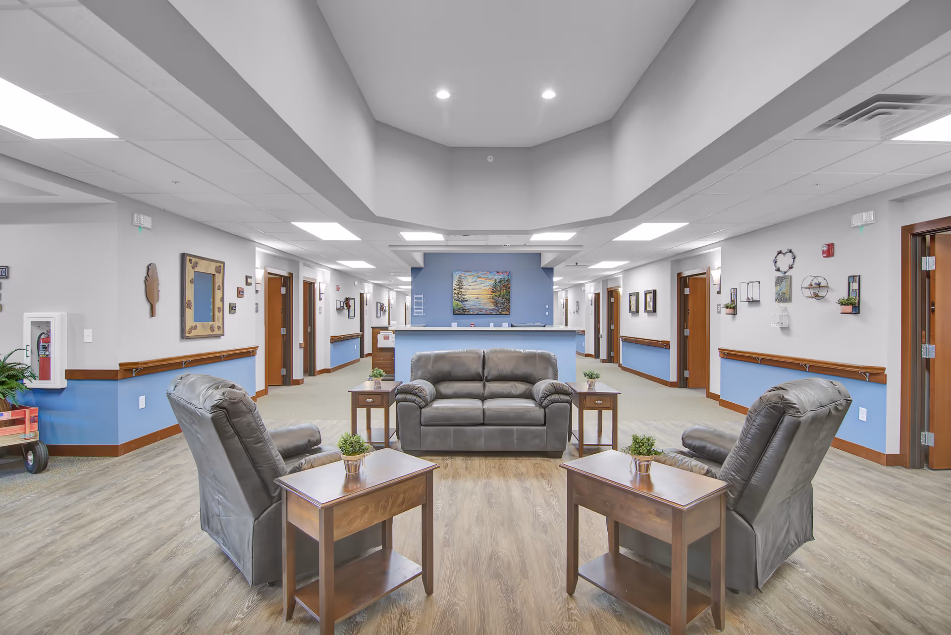 A spacious senior living facility common area with two gray leather armchairs and a matching loveseat arranged around wooden side tables with small potted plants. The room has light wood flooring, light gray walls with blue wainscoting, and a blue accent wall with a colorful painting. Hallways with multiple doors extend on both sides, and the ceiling has recessed lighting.