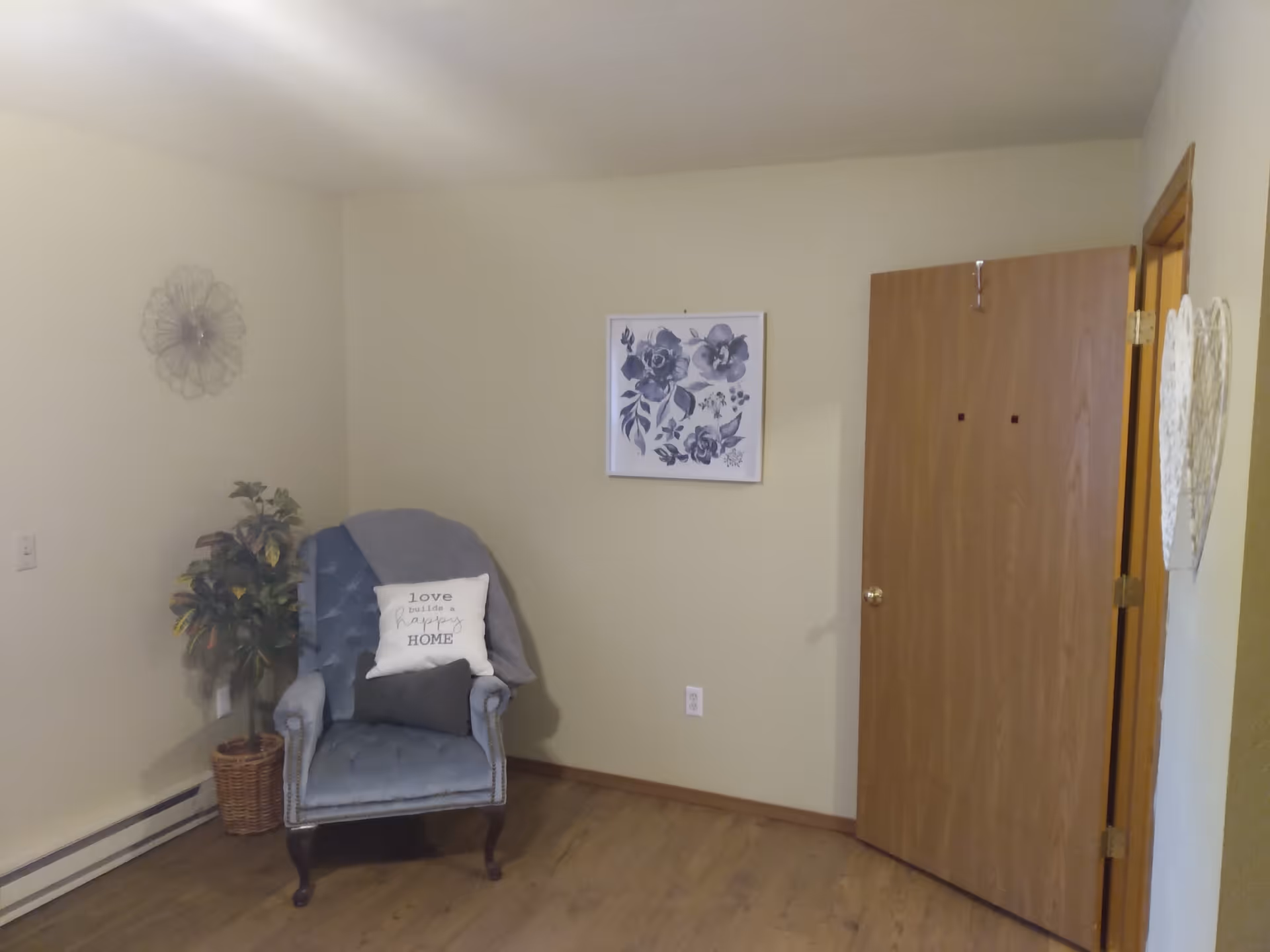 A cozy corner of a room with a blue upholstered armchair that has a white pillow with the text 'love builds a happy HOME' and a smaller dark pillow. A gray blanket is draped over the back of the chair. Next to the chair is a potted plant in a wicker basket. On the wall above the chair is a framed floral artwork in shades of blue. To the right, a wooden door is partially open, and a white decorative heart hangs on the adjacent wall. The room has light-colored walls and wood flooring.