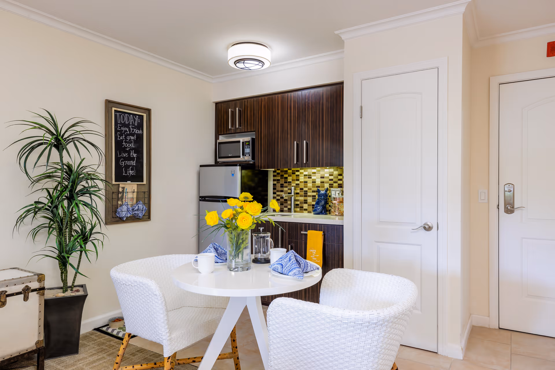 A small kitchen and dining area with a round white table set with blue napkins, white cups, and a vase of yellow flowers. The kitchen features dark wood cabinets, a microwave, a refrigerator, and a backsplash with yellow and brown tiles. There is a tall green plant in a black pot and a framed chalkboard on the wall with the message: 'TODAY: Enjoy Friends, Eat great food, Live the grand Life.' Two white woven chairs are placed around the table. Two white doors are visible on the right side of the image.