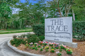 Stone sign for The Trace Assisted Independent Living surrounded by a landscaped flower bed with pink and white flowers, green shrubs, and trees in the background under a clear sky.