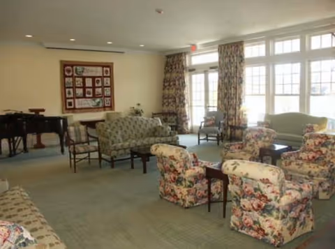 A spacious living room with multiple floral-patterned armchairs and sofas arranged around small wooden tables. Large windows with floral curtains allow natural light to fill the room. A grand piano is visible in the corner, and a quilted wall hanging decorates one wall.