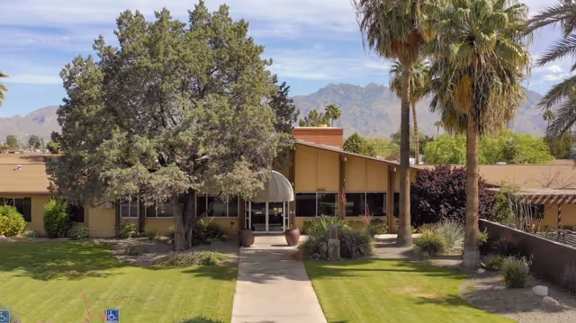 Front entrance of a one-story rehabilitation center with a sidewalk, green lawn, palm trees and mountains in the background.