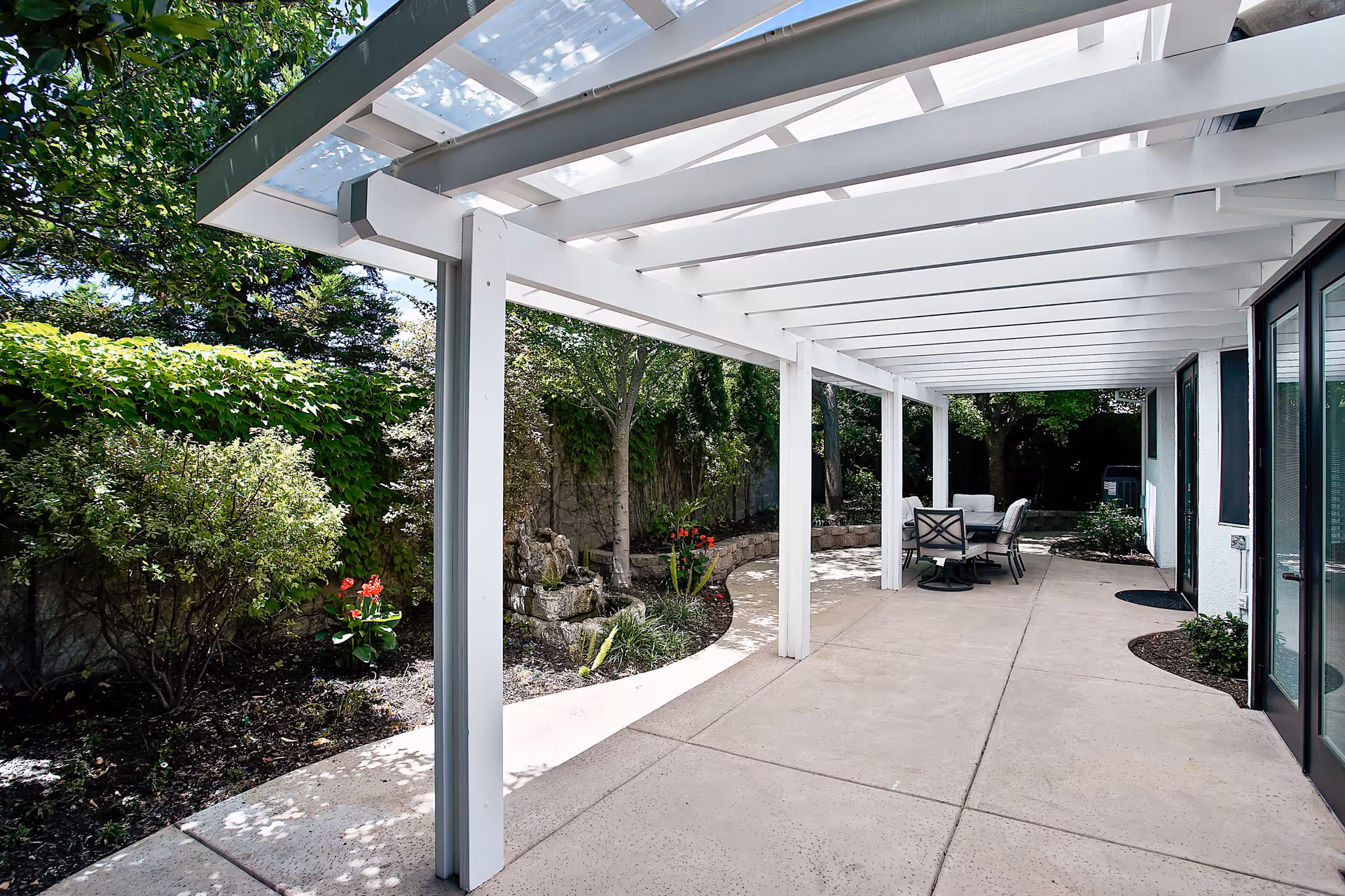 Outdoor patio area with a white pergola overhead, concrete flooring, and a round table with chairs. Surrounding the patio are green bushes, trees, and a stone retaining wall with some red flowers.
